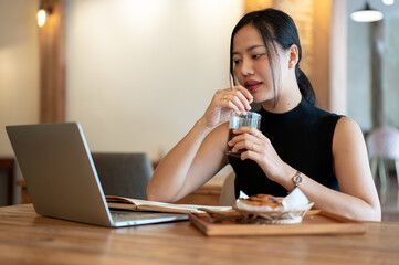 A woman is sipping coffee while looking at her laptop screen, working remotely at a coffee shop.