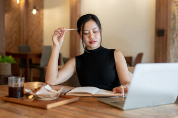 A thoughtful Asian woman focuses reading a book and working on her tasks at a coffee shop.