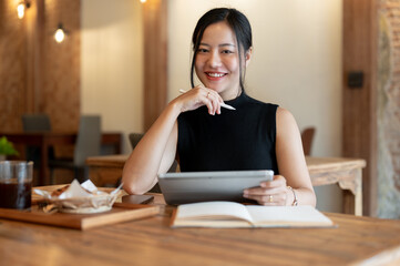 A beautiful Asian woman is smiling at the camera while sitting in a coffee shop with her tablet.