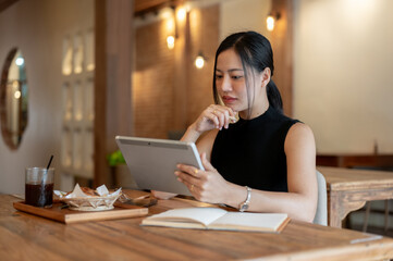 A thoughtful and gorgeous Asian woman using her digital tablet while sitting in a beautiful cafe.