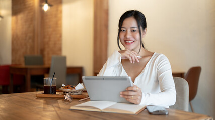 A beautiful Asian woman sits at a table in a beautiful coffee shop with a digital tablet in her hand