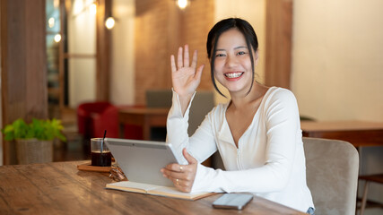 A cheerful woman sits at a table in a coffee shop, holding her tablet, waving her hand at the camera