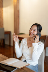 A cheerful Asian woman enjoys talking on the phone with her friend while relaxing in a coffee shop.