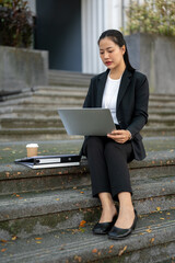 Busy, concentrated Asian businesswoman is working on her laptop while sitting on stairs in the city.