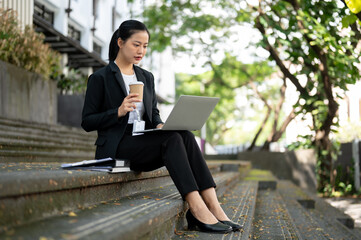 A busy Asian businesswoman is sipping coffee and working on her laptop while sitting on stairs.