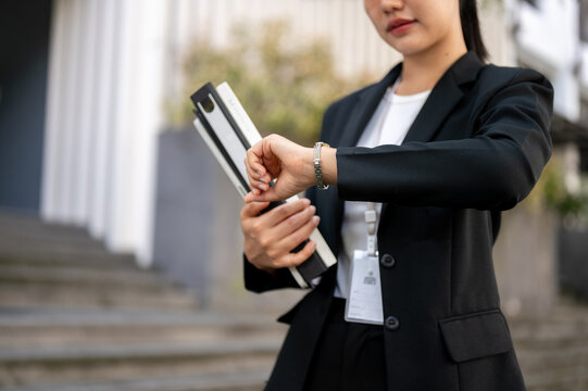A busy Asian businesswoman is checking time on her wristwatch while walking in the city street. - Powered by Adobe