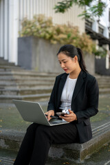 A professional Asian businesswoman is working on her laptop while sitting on the stairs in the city.