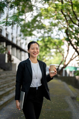 A confident Asian businesswoman is walking down the stairs with a coffee cup and laptop in her hands