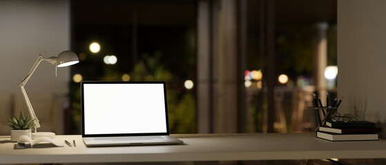 A white-screen laptop mockup, a table lamp, and accessories on a table in a modern room at night.