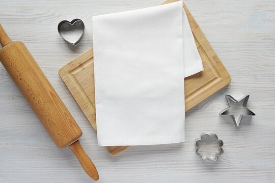 White plain kitchen towel mockup for design or text presentation, wooden cutting board, kitchen utensils, natural flat lay composition on light color wooden table.
