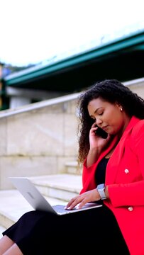 Black businesswoman working on laptop while talking on phone outdoors. Curvy woman sitting on stairs.