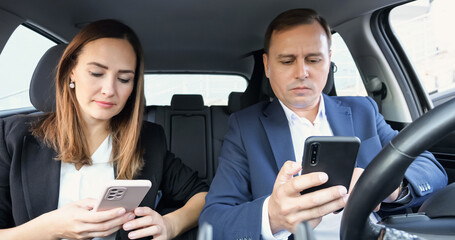 Office workers man and woman in suits sit in car holding phones. Colleagues check social media news talking. Businesswoman smiles reading messages on phone