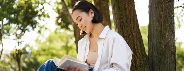 Beautiful smiling girl reading book in park on sunny day, sitting near tree on lawn and relaxing outdoors