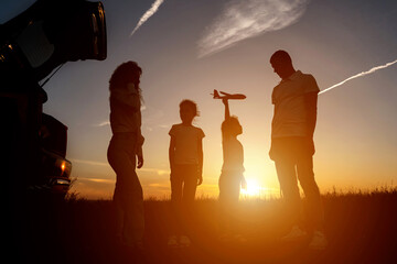 Black silhouettes of family members fly kite near car with open trunk in evening, sunlight. Concept...