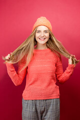 Healthy lifestyle treatment cold season concept. One Smiling Happy Cheerful Woman Wear Warm Coral Sweater and Hat While Gesture With Hair on Plain Red Background