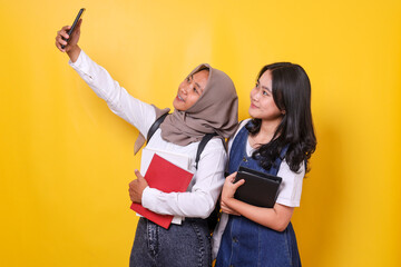 Cheerful student girls holding book and do selfie on mobile phone over yellow background