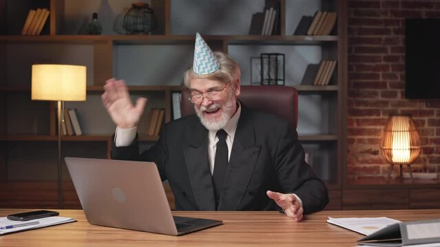 Excited businessman about 60 wearing formal attire and birthday hat holding virtual conference on laptop in workplace. Happy bearded entrepreneur enjoying compliments on successful startup online.
