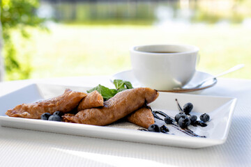 Close-up a white cup of coffee with pancakes and fruit on the white plate is on the table against the backdrop of the river. Tea party in nature. Coffee morning