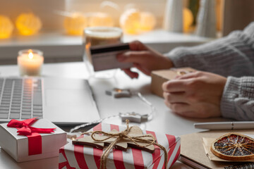 Woman using laptop and credit card, making online purchases at cozy home. Holiday shopping. Gift present box, decoration. Winter sales, Black Friday. Christmas discount promotions. Blurred bokeh light