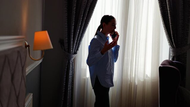 Angry woman call phone at hotel room during business trip. Annoyed person talking mobile phone at window apartment. Upset woman using cellphone. Businesswoman arguing in phone conversation silhouette.