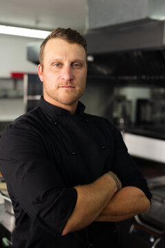 Confident caucasian male head chef in black uniform with arms crossed standing in kitchen