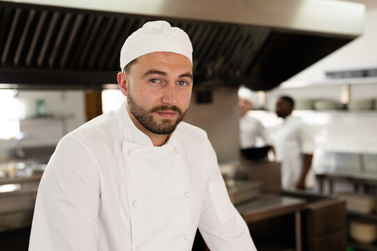 Portrait of confident caucasian male chef wearing hat standing in kitchen at cooking school