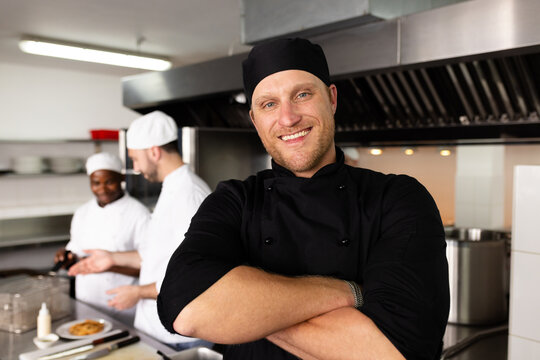 Diverse smiling male head chef with arms crossed standing against students cooking in background
