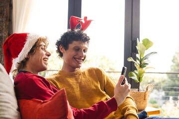 Happy diverse gay male couple embracing wearing christmas hats and having video call at home