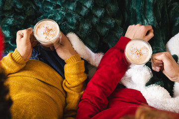 Diverse gay male couple holding glasses of cocoa covered with blanket at home
