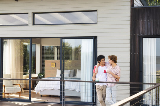 Happy diverse gay male couple embracing and holding mugs of coffee at balcony