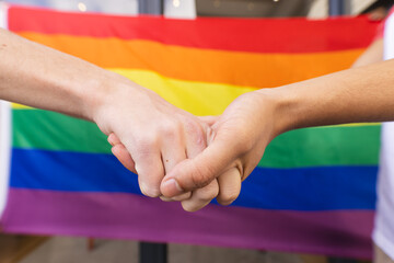Diverse gay male couple holding lgbt flag and hands at balcony