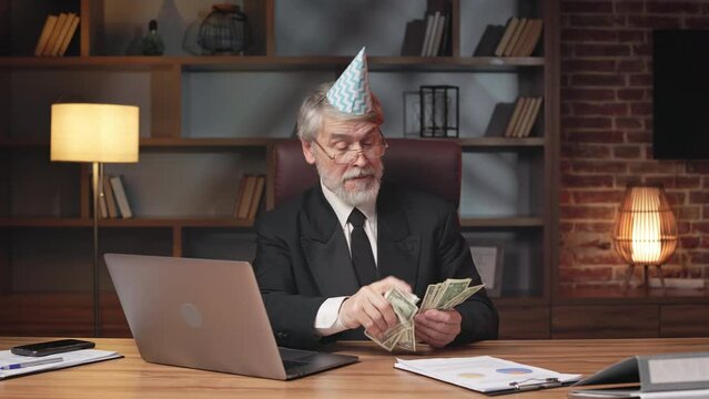 Displeased older man in paper cone hat thumbing through cash before leaving it on wooden desk in office. Upset business person feeling stress while struggling with money problems in workplace.