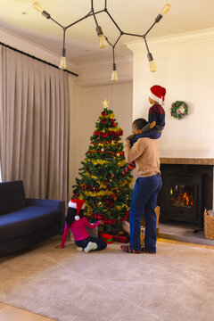 African american father, son and daughter decorating christmas tree at home, copy space