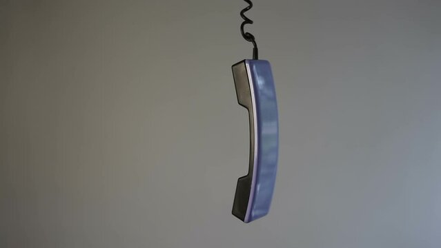 An abandoned landline telephone handset hangs on a cord on a gray background. The telephone receiver is hanging and dangling in the air. Unfinished phone conversation.