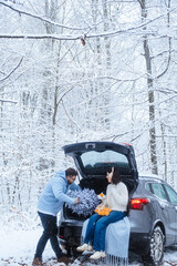 In a winter forest, a woman sits in a car trunk with a net of tangerines, and a man carries a tree