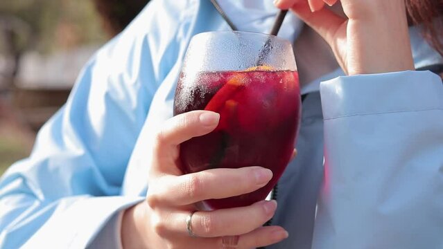A girl in an autumn raincoat drinks sangria against the backdrop of a garden, an open-air cafe. Female hands with a glass close-up. Glass of sangria