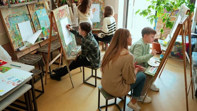 Top view of concentrated young children talking and painting during an illustration class with teacher at art school