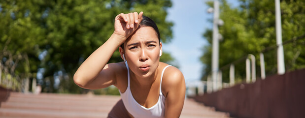 Portrait of asian woman taking break, breathing heavily and panting after running, jogger standing...