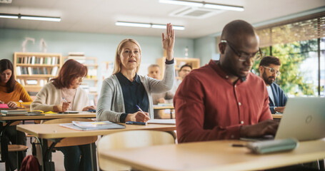 Diverse Mature Students Learning in Classroom, Using Laptops and Writing in Notebooks. Female Raising Hand and Asking Teacher a Question During a Lecture in Adult Education Course in School