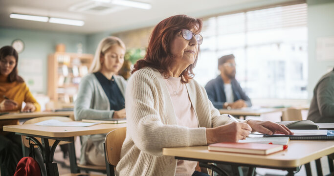 Portrait of a Happy Senior Woman Taking a Course in an International Adult Education Center. An Alderly Female Wearing Glasses, Sitting Behind a Desk, and Writing Down Notes in a Notebook.