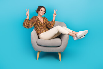 Full length photo of adorable cheerful person sit cozy chair hands fingers demonstrate v-sign...