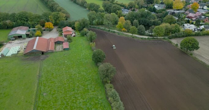 4k 60fps drone footage of a tractor ploughing through a brown field with a village in the background