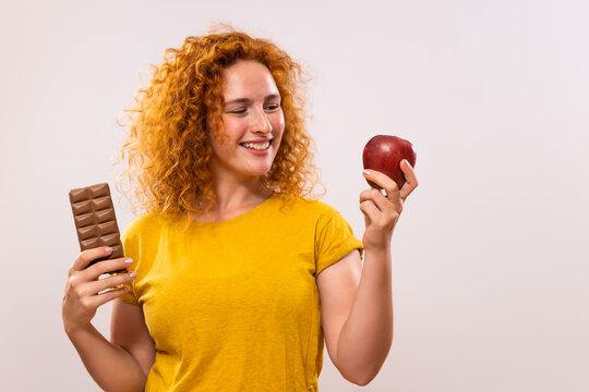 Beautiful ginger woman is holding apple and chocolate. She has  decided to eat healthy.