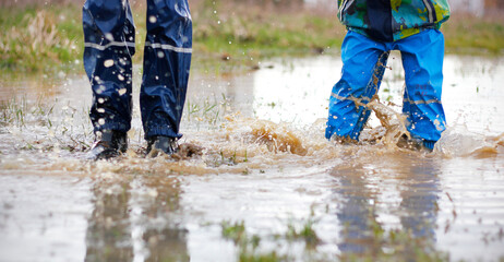 Legs of two boys wearing blue waterproof pants and rubber boots jumping in a muddy puddle during an autumn or spring walk. Outdoor fun in the rainy weather. Health and immunity of children concept - obrazy, fototapety, plakaty