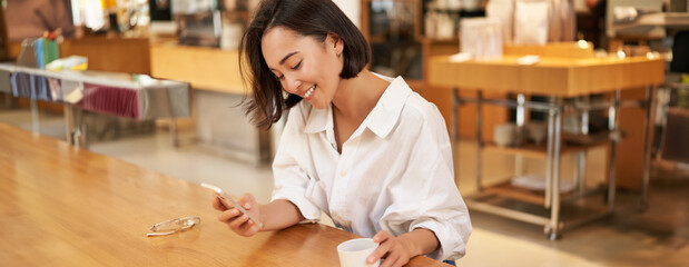 Portrait of beautiful asian woman with smartphone, relaxing in cafe, sitting and enjoying coffee...