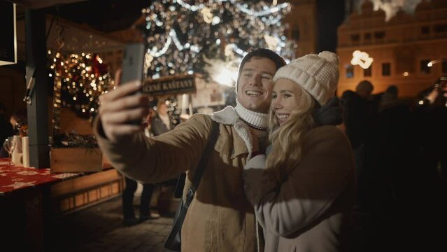 Caucasian couple or family having a video call with friends family, selfie Christmas New Year holidays at illuminated background of decorated town square. Woman and man in love enjoy winter vacation