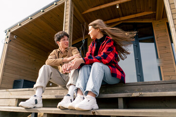 Fototapeta na wymiar A young stylish couple in love sits together hugging on the steps of large wooden cottage and chats cheerfully