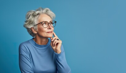 Senior woman with a finger tip on her cheek, thinking, happy with herself and her life. Best-ager, baby-boomer with grey short hair on a blue background.