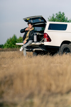 The open trunk of an SUV parked in forest-steppe Guy sits squinting in the sun and drinks coffee