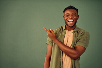 Choose this. African american male person with toothy smile on face pointing with index finger on empty space over green background. Handsome young guy indicating place for advertising text in studio. - obrazy, fototapety, plakaty